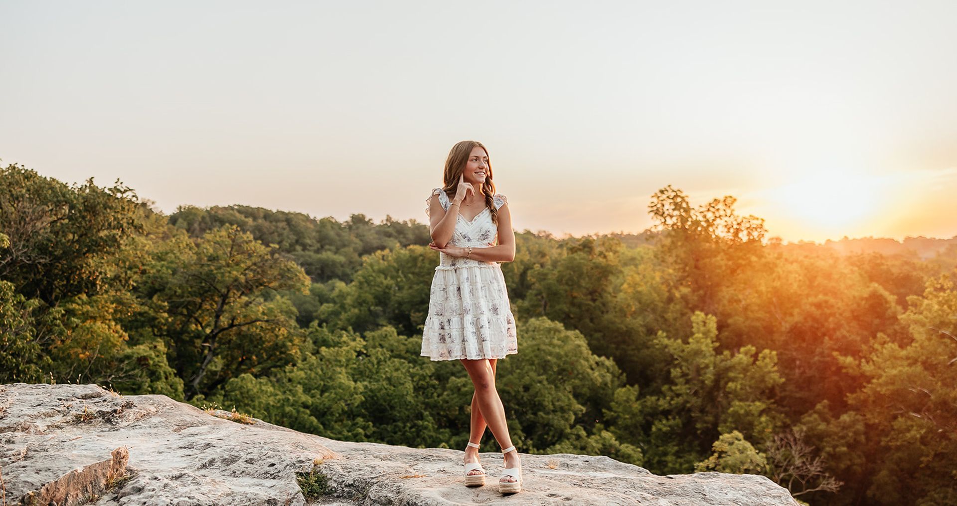Woman in a white dress standing on a cliff, looking up at the sunset over a forest.