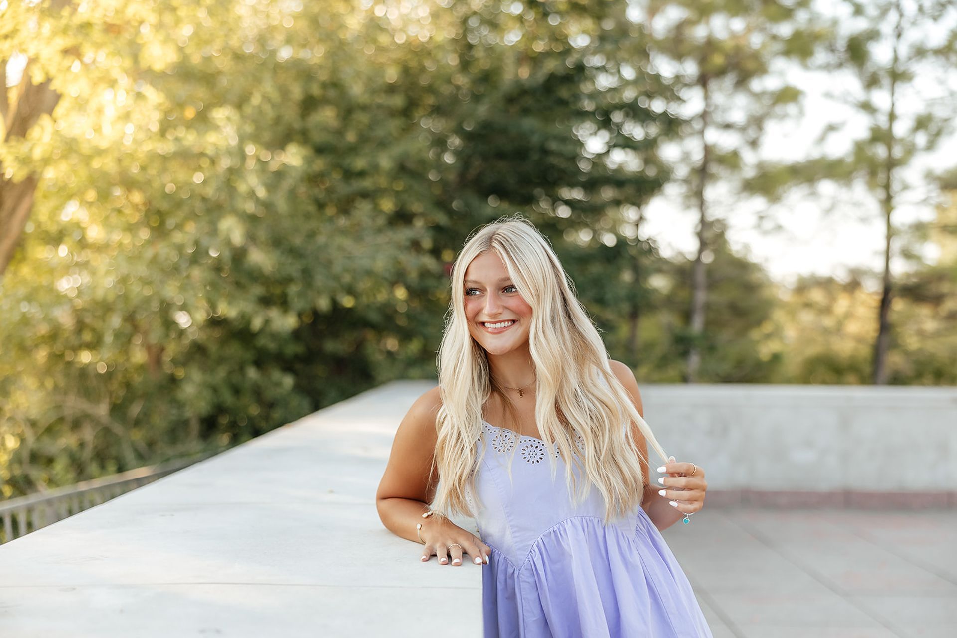Blonde-haired person in a purple dress smiles, leans on a ledge, outdoor setting.