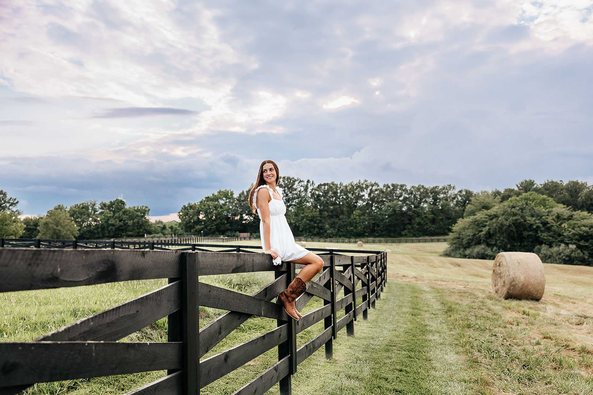 Woman in white dress and boots sits on a black fence, field and sky background.