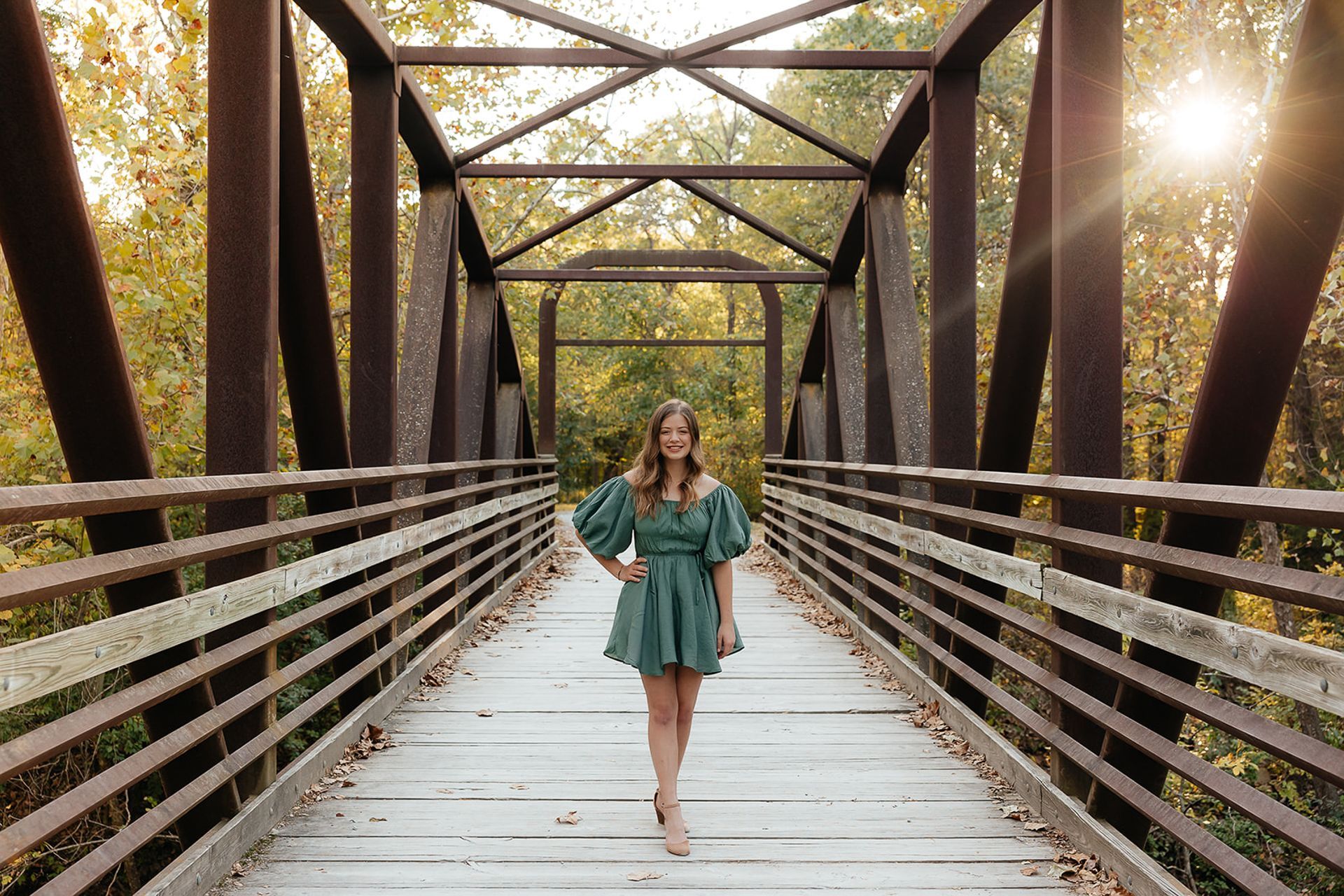 Woman in a green dress stands on a bridge, smiling. Fall foliage surrounds the bridge with sunlight.
