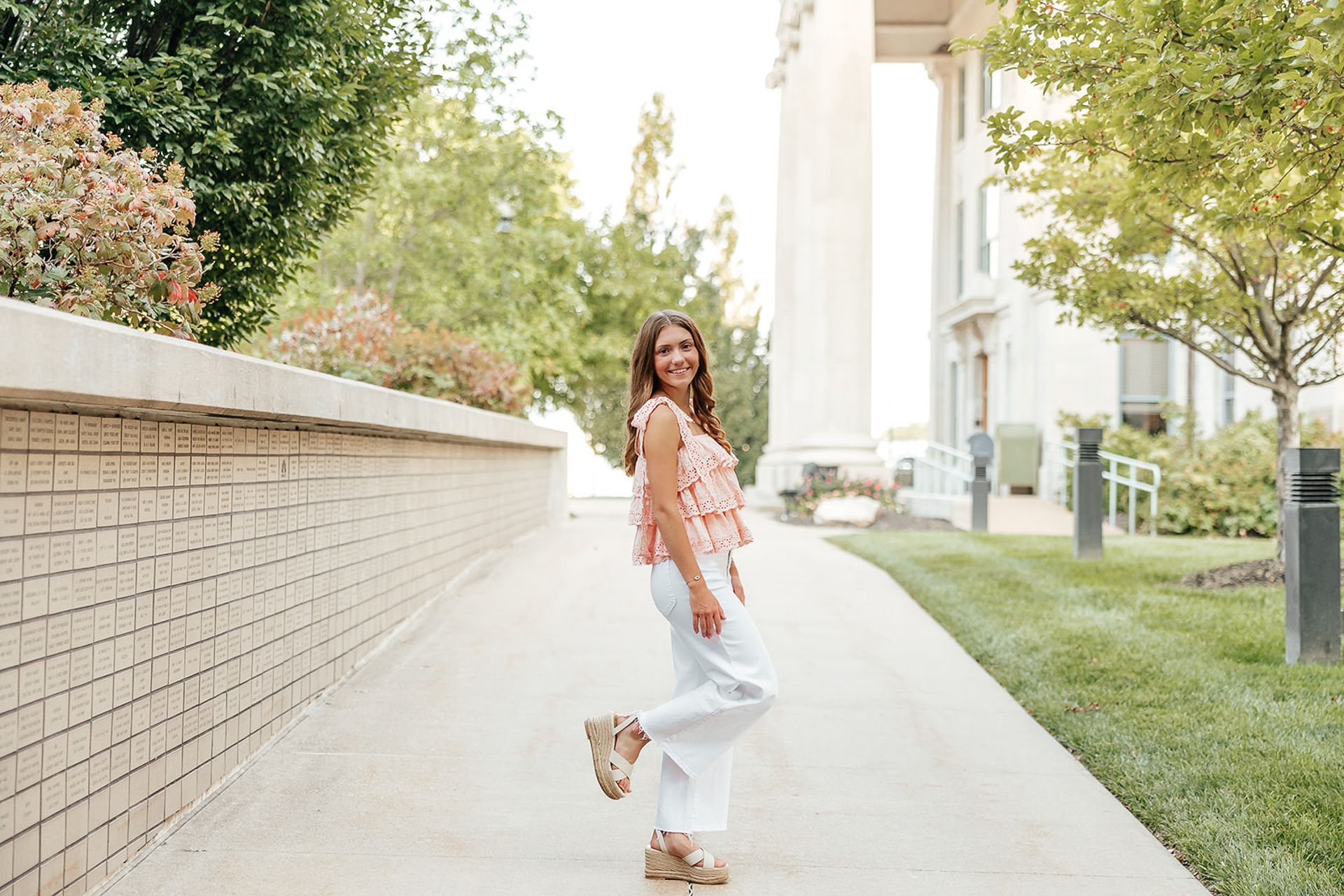 Woman smiles, poses with one foot up on sidewalk, near brick wall and building.