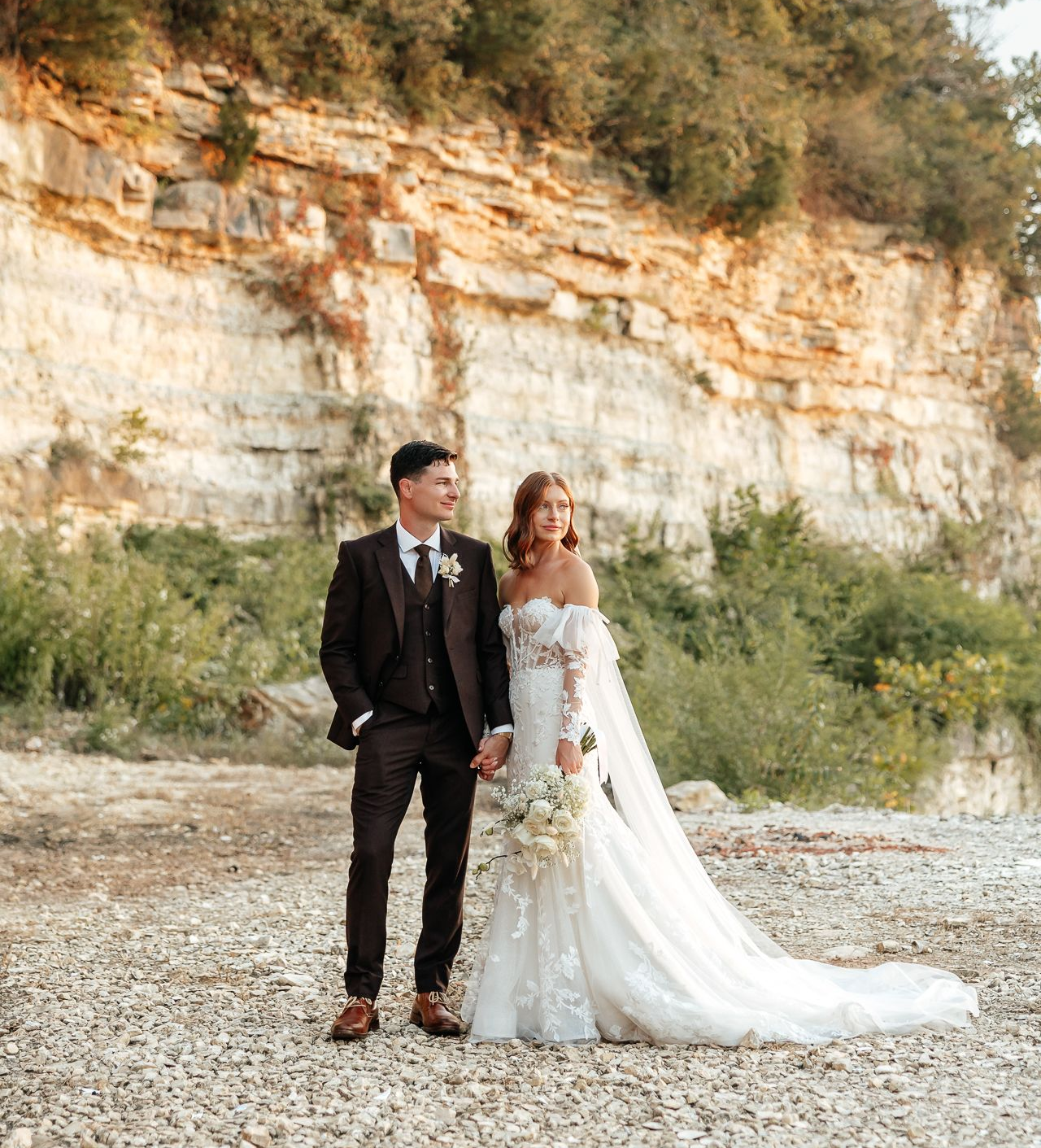 Couple in wedding attire holding hands, posing in front of a rocky cliff.