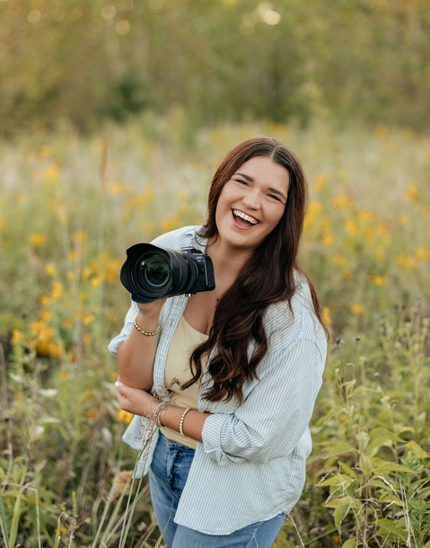 Woman holding a camera, smiling widely in a field with yellow flowers. Wearing a light blue shirt, yellow top, and jeans.