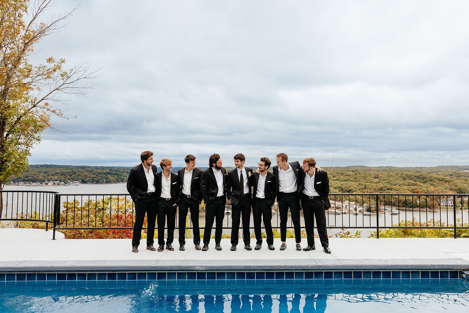 Men in black suits stand by a pool overlooking a lake under a cloudy sky.