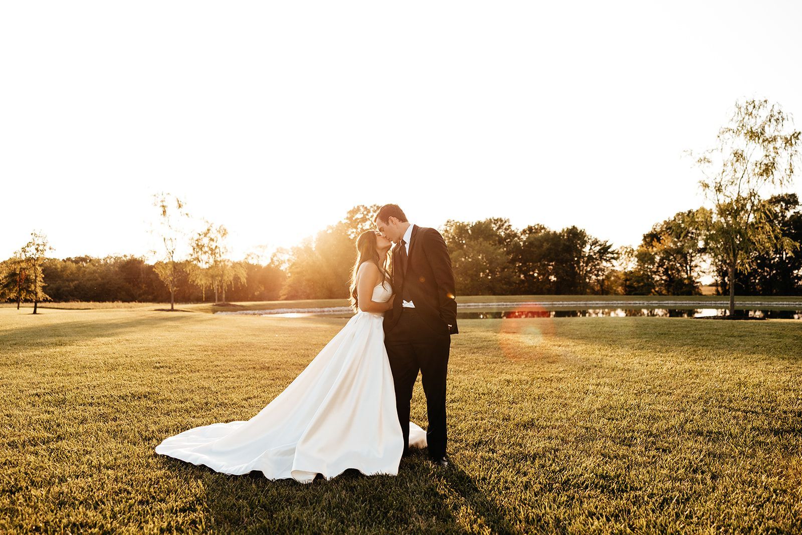 Couple kissing in a field at sunset; bride in white gown, groom in a black suit.