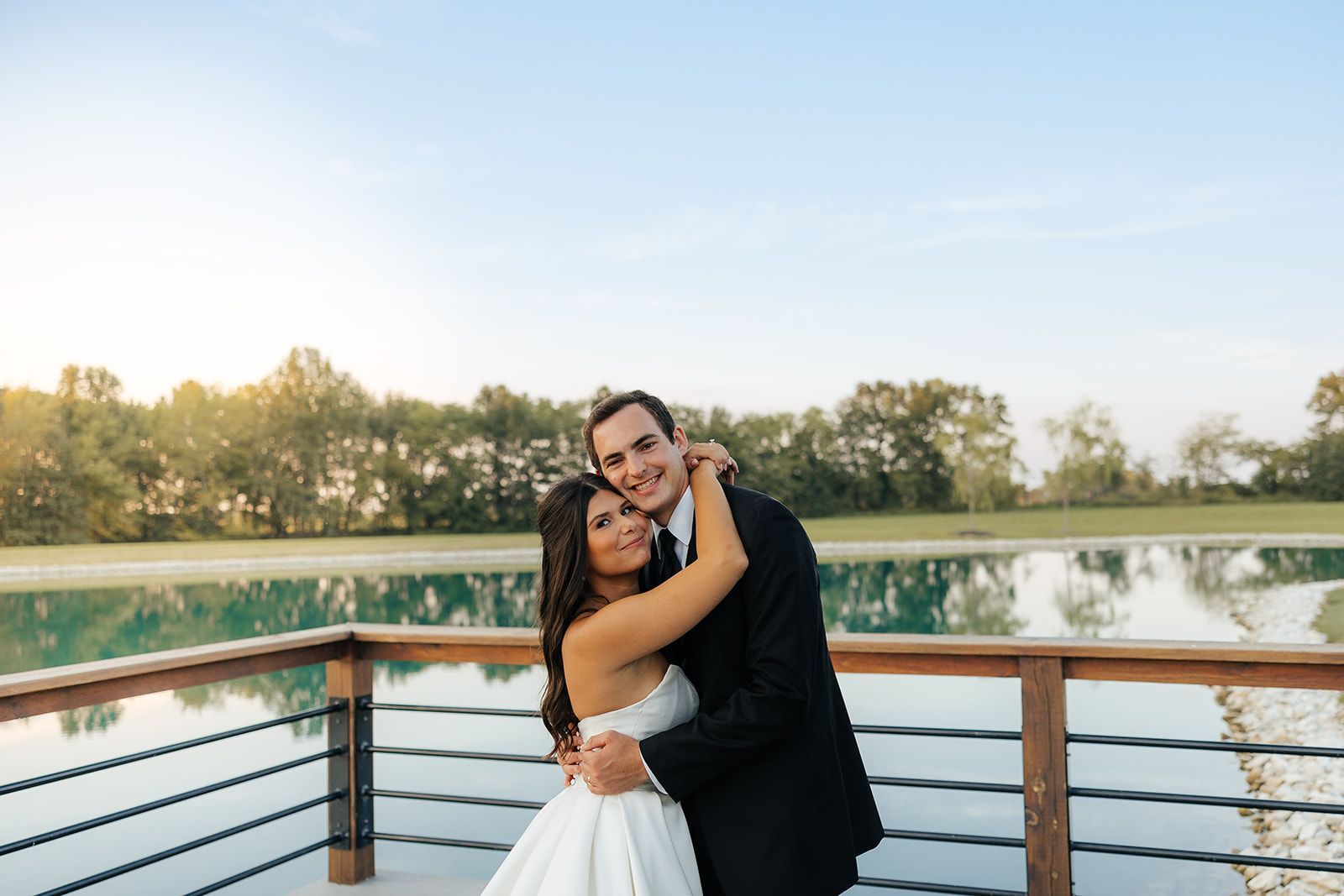 Bride and groom embrace on a wooden deck overlooking a lake, sunny sky.