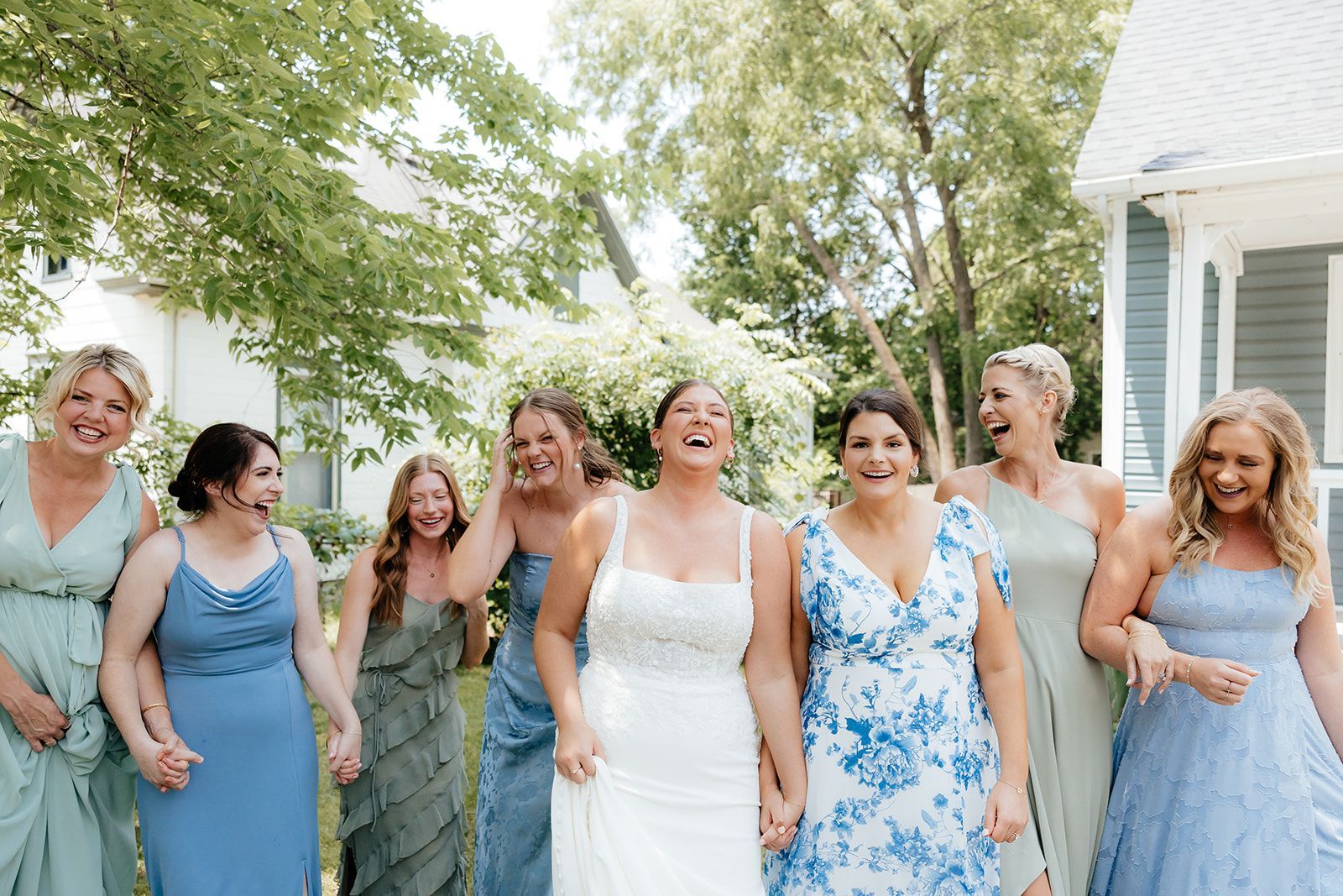 A bride in a white dress laughs with bridesmaids in blue and green dresses outdoors.