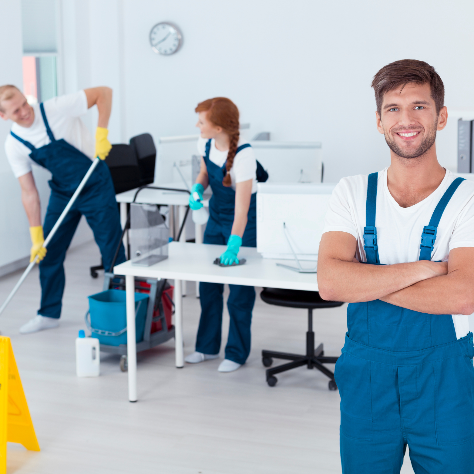 A man in blue overalls is standing in an office with his arms crossed