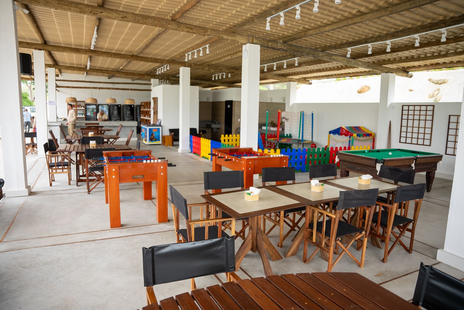 Área de lazer coberta com mesas, pebolim, mesa de sinuca e área de recreação infantil, sob um teto de bambu.