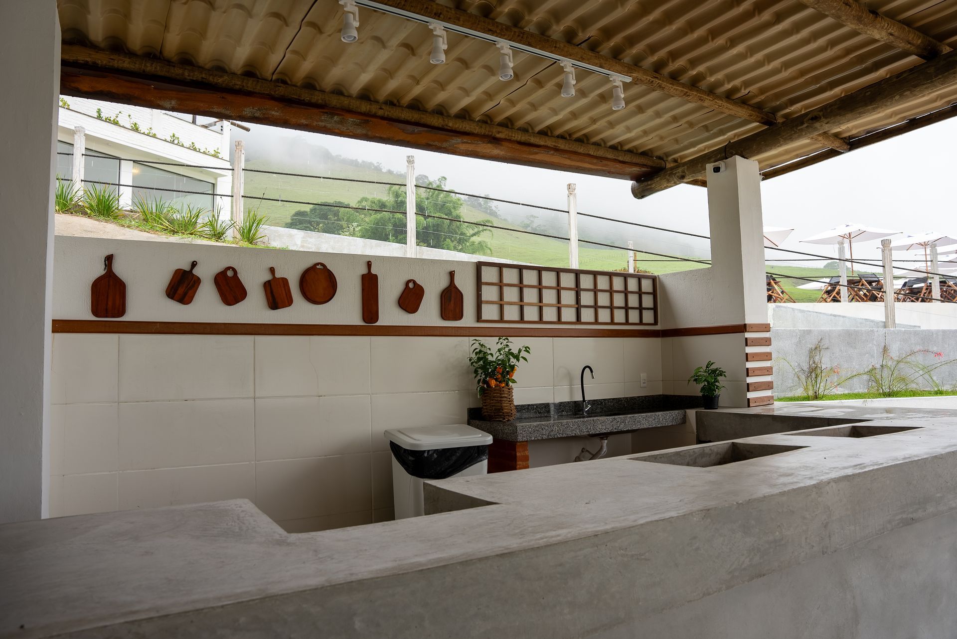Cozinha ao ar livre com bancada de concreto, pia e tábuas de corte decorativas sob um pátio coberto.
