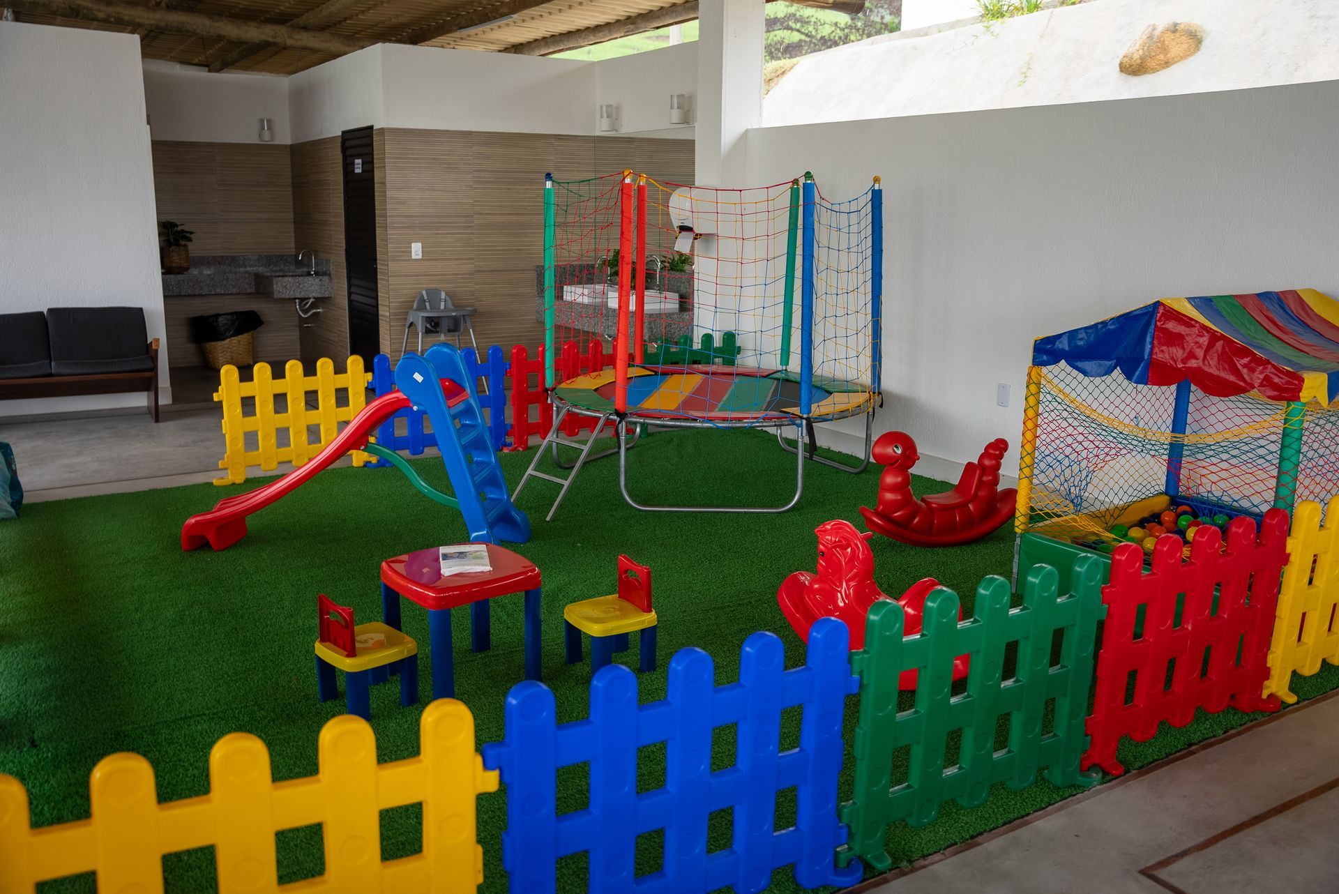 Área de lazer com escorregador, cama elástica, piscina de bolinhas e cerca colorida em grama sintética.