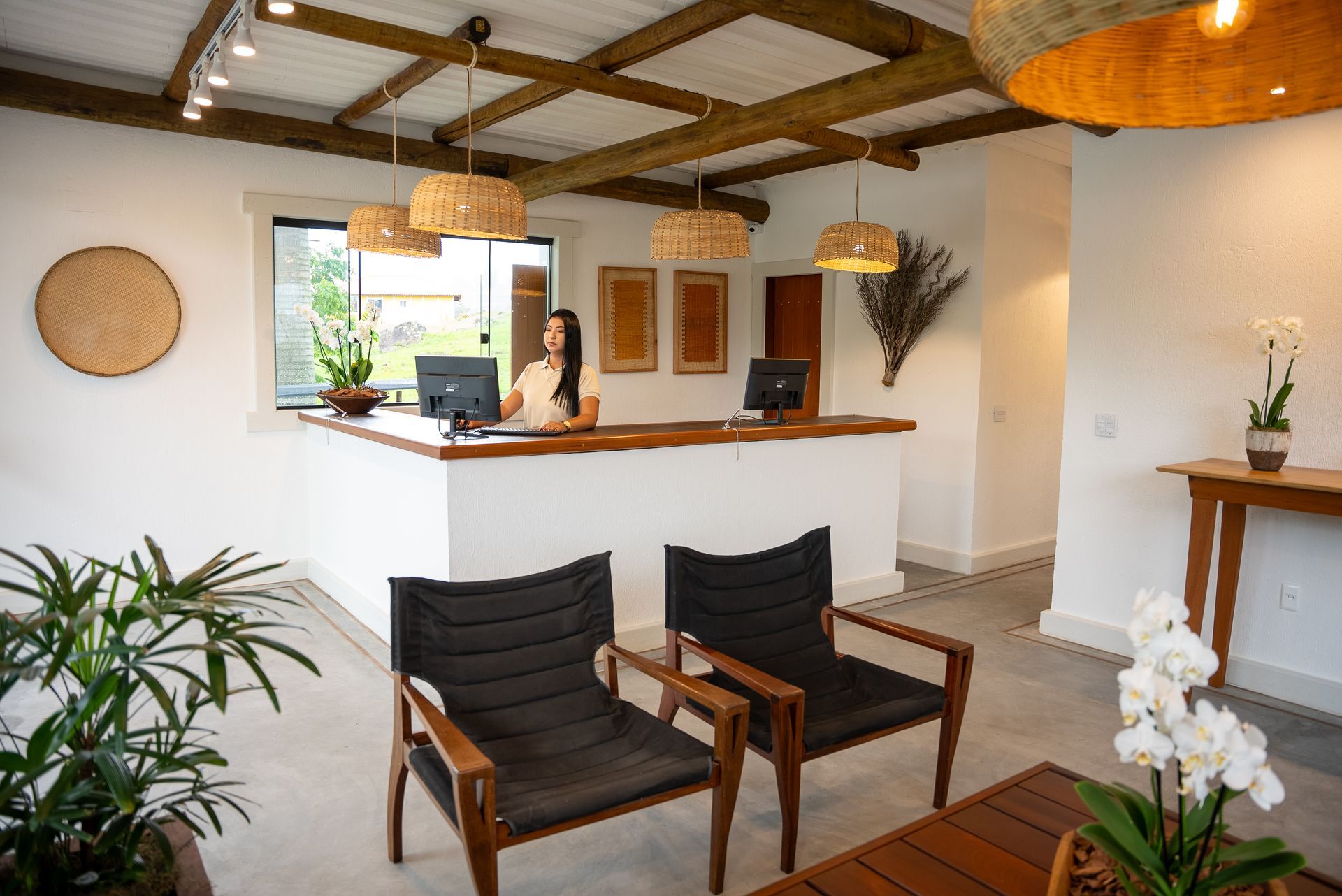 Área de recepção com uma mulher no balcão, duas cadeiras pretas e decoração em tons naturais.