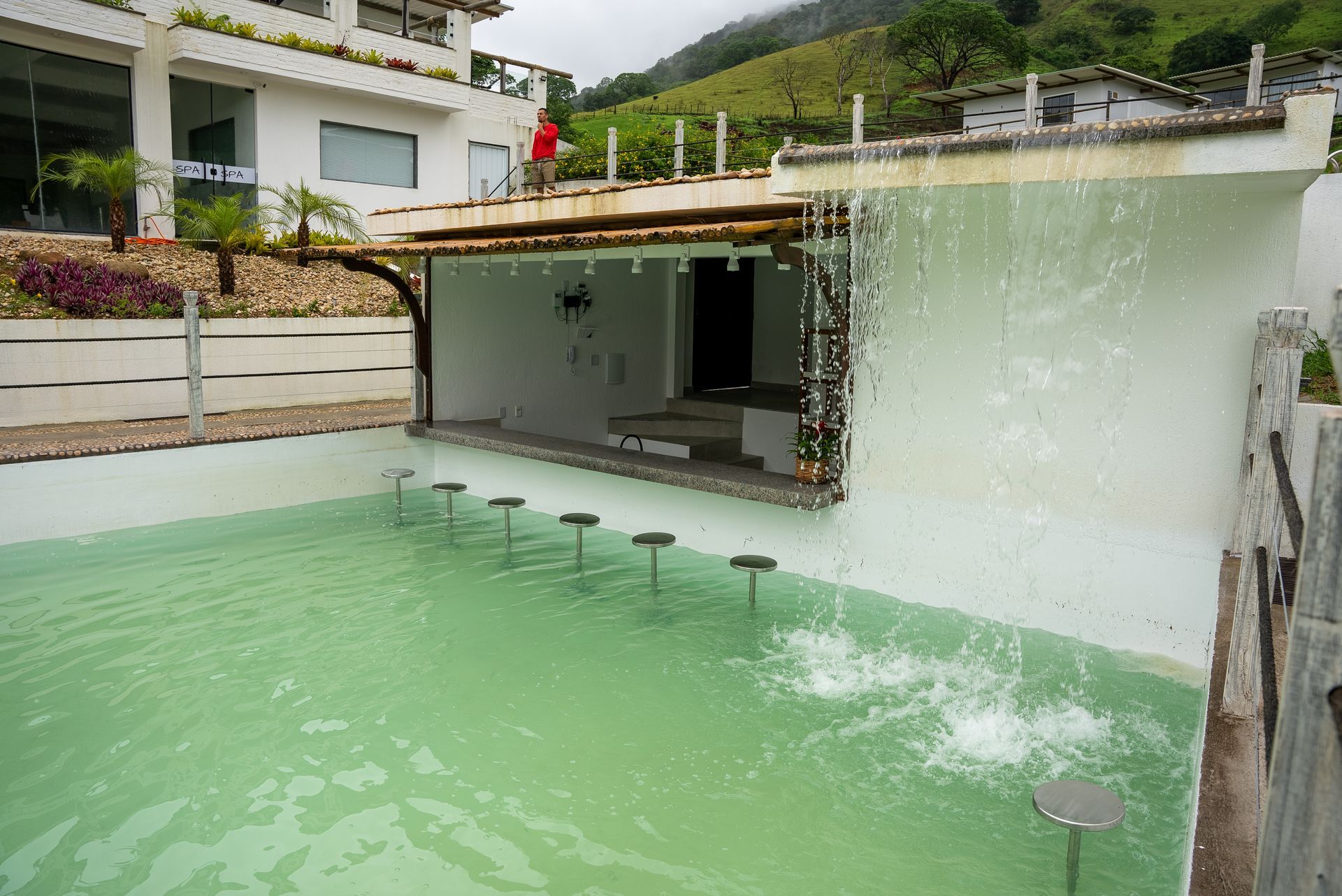 Piscina com cascata e bar ao lado, em frente a um prédio branco e montanhas ao fundo.