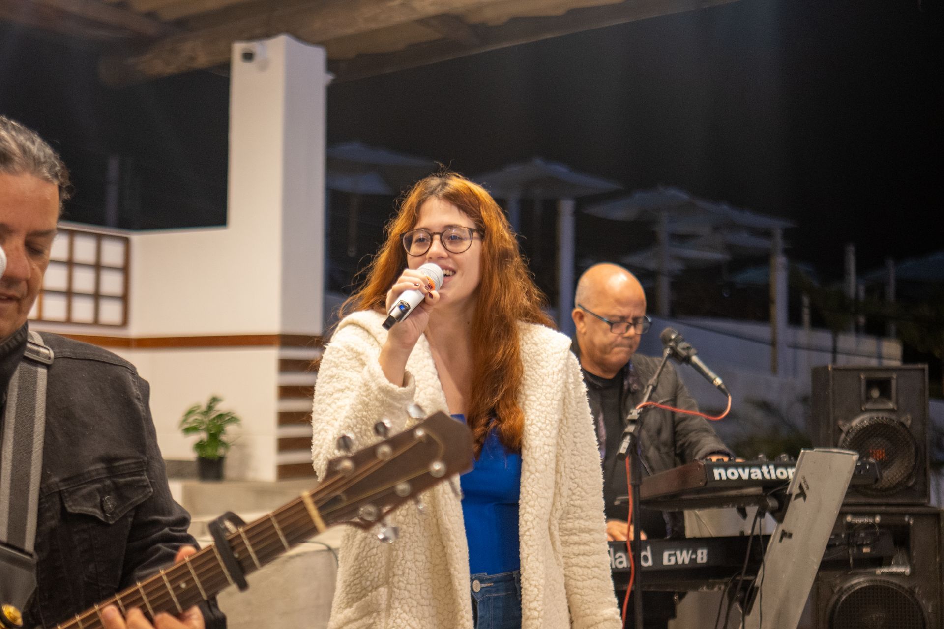 Uma banda se apresentando ao ar livre à noite: uma mulher cantando em um microfone, um guitarrista e um tecladista.