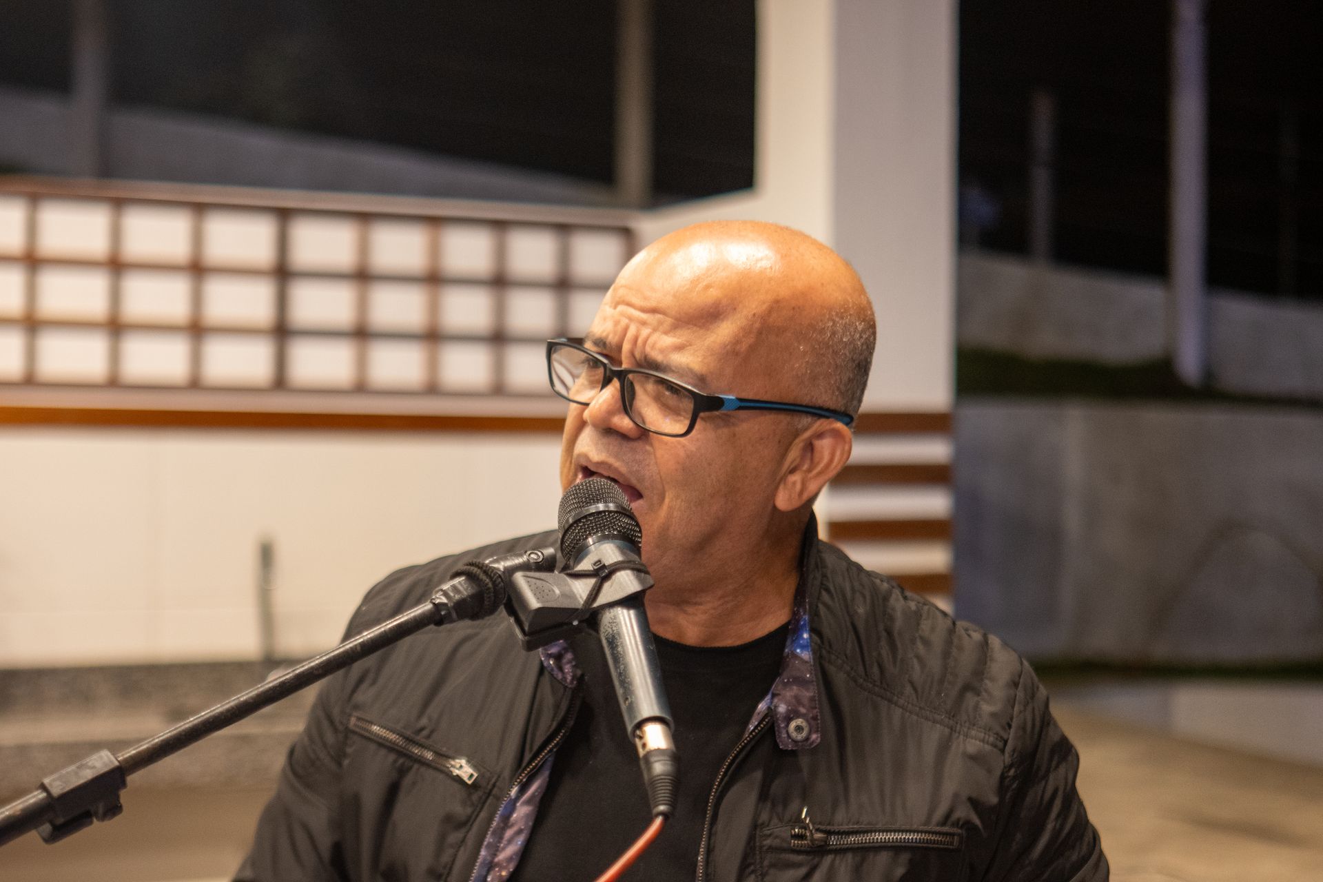 Homem de óculos canta em um microfone, vestindo uma jaqueta preta, em um ambiente interno.