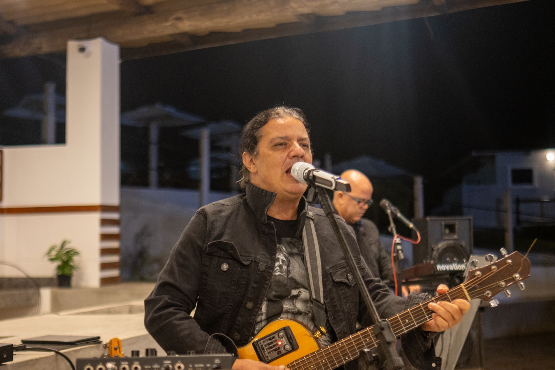 Homem cantando e tocando violão em um palco à noite, com outro músico ao fundo.
