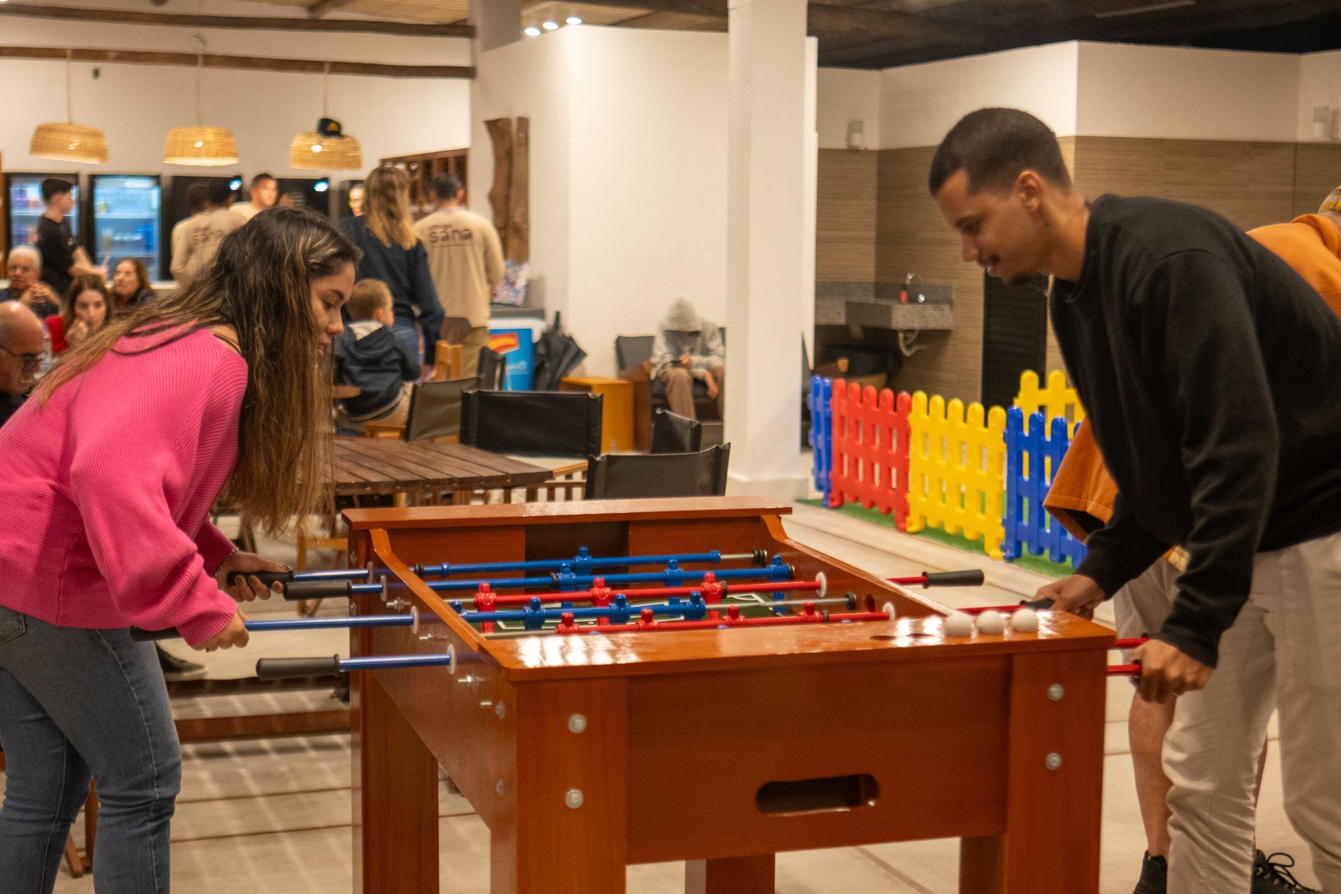 Duas pessoas jogam pebolim em uma sala bem iluminada. Uma cerca colorida as separa de uma área com uma pia.