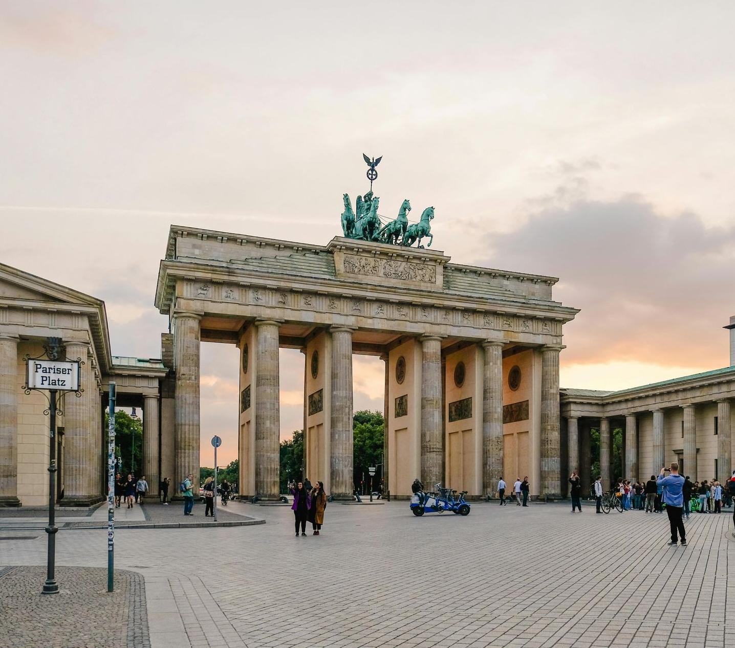 Brandenburg Gate, Berlin, with columns, chariot sculpture on top, people in plaza, cloudy sky.
