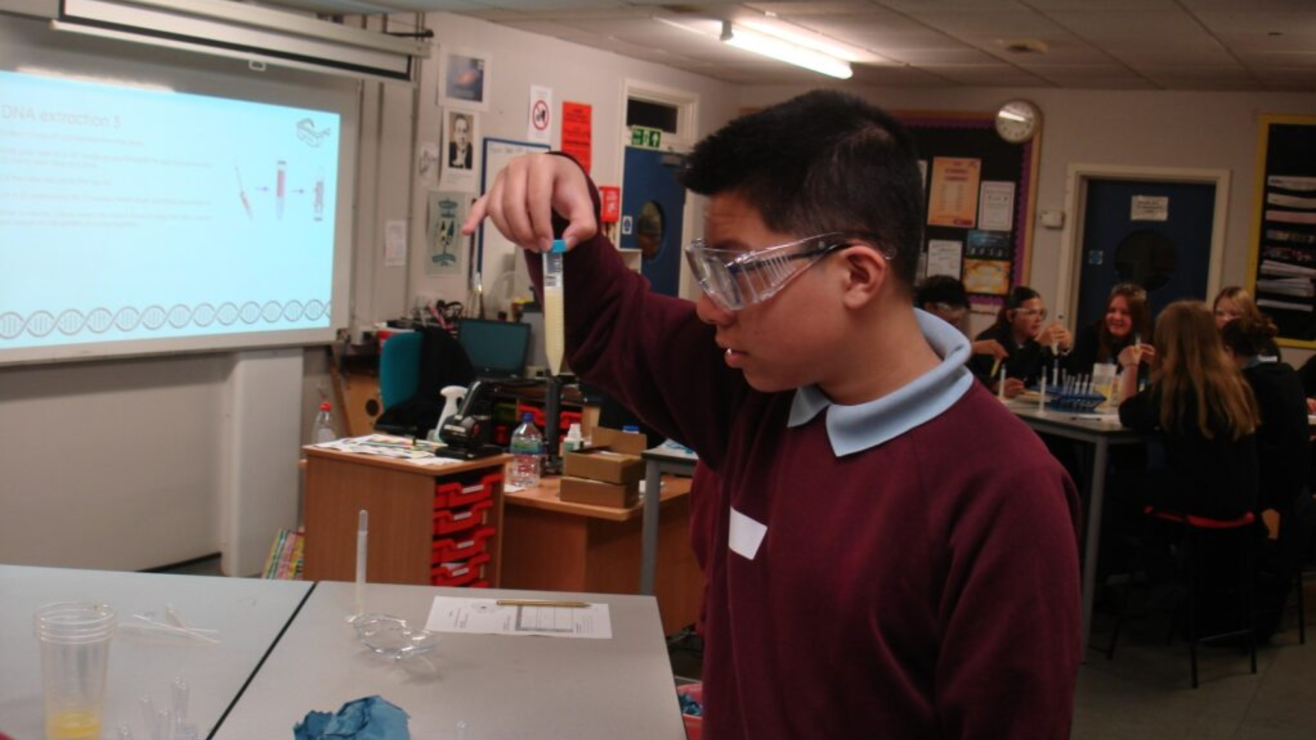 Student in lab coat and goggles holding a test tube with liquid during a science experiment in a classroom.