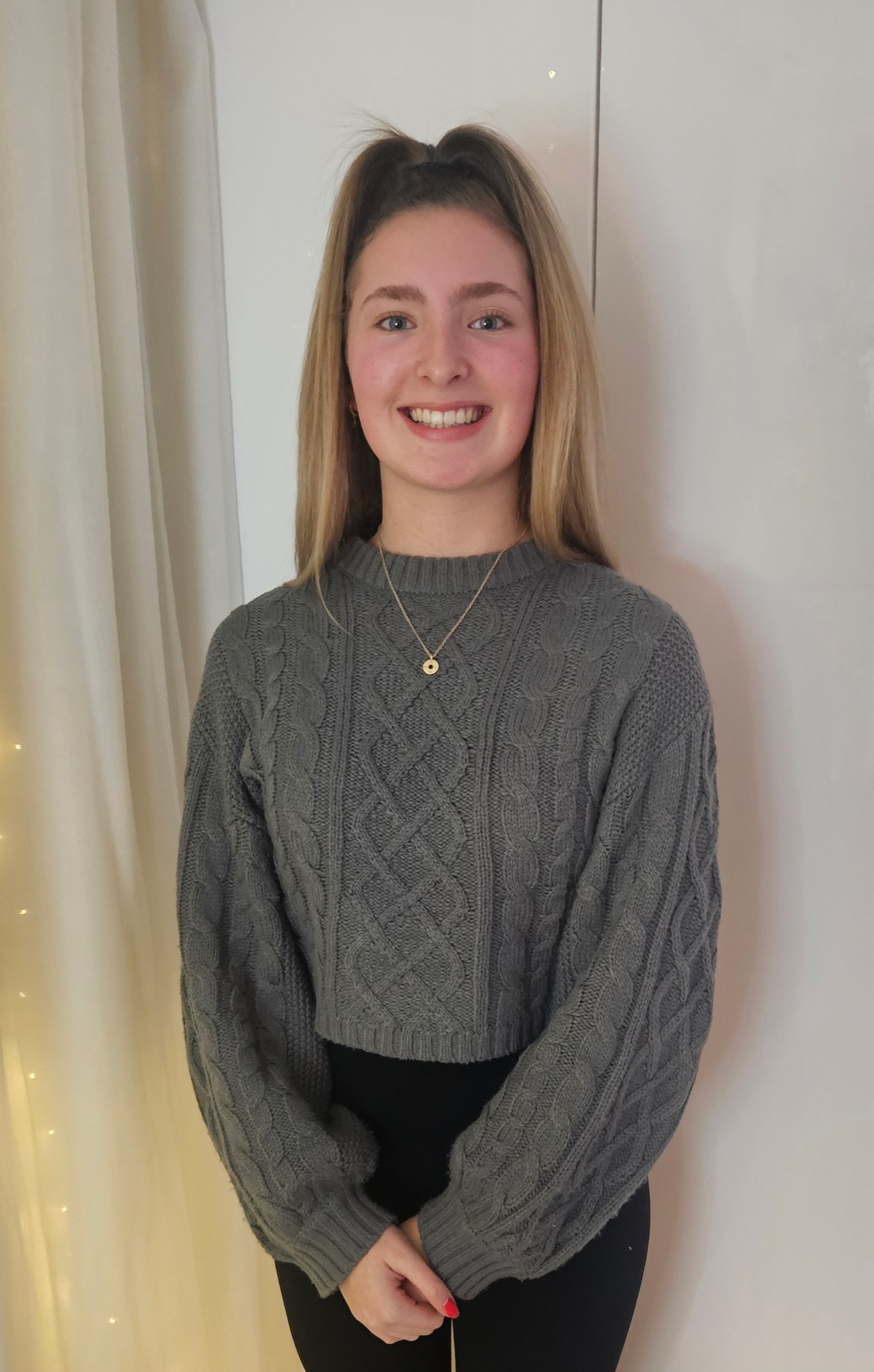 A young woman in a grey sweater and black pants is smiling for the camera.