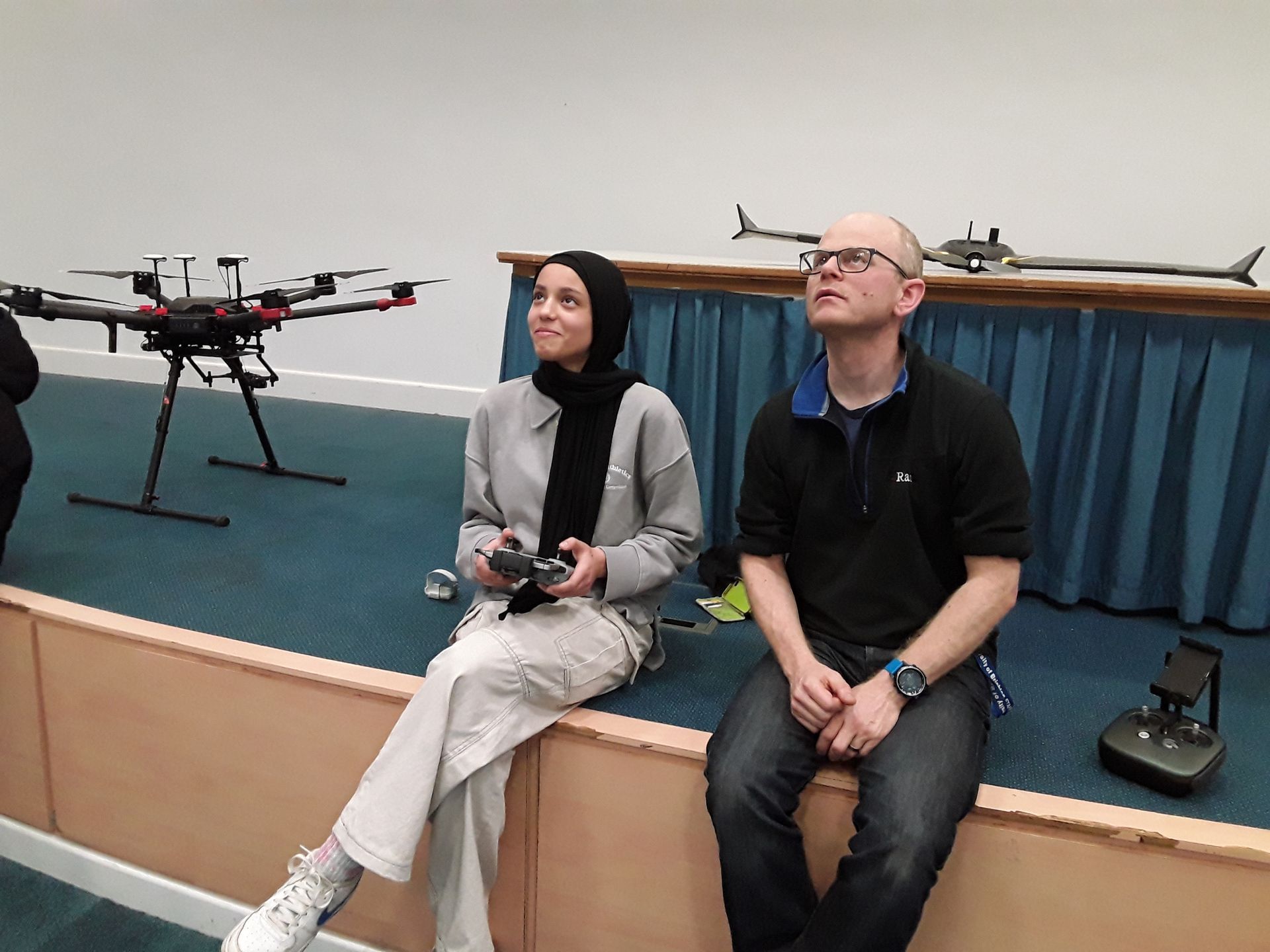 A man and a woman are sitting on a stage looking up at a drone.