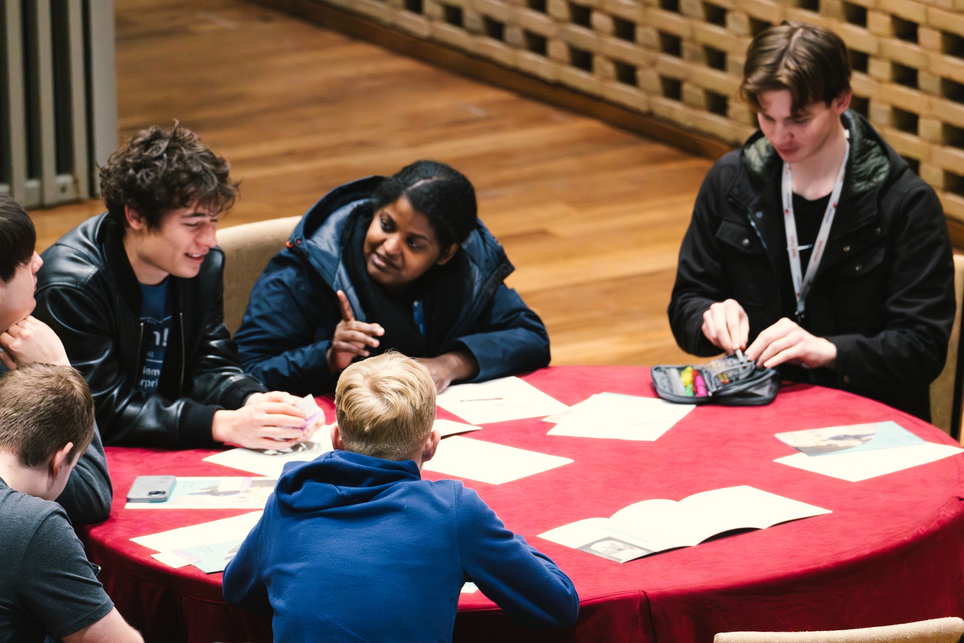 People seated around a red table, working on papers. Two are smiling and talking. Indoors.