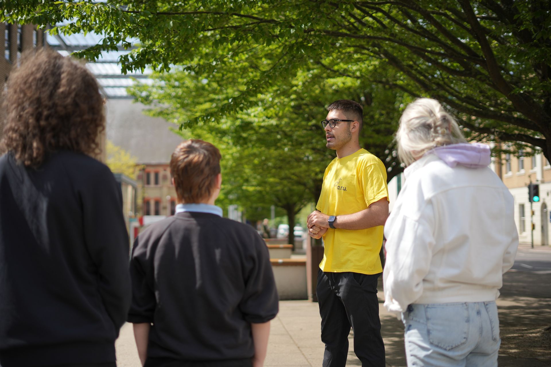 A man in a yellow shirt is talking to a group of people on a sidewalk.