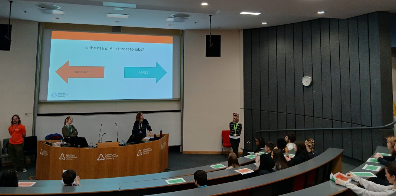 A lecture hall presentation with presenters on the stage and an audience in curved rows.