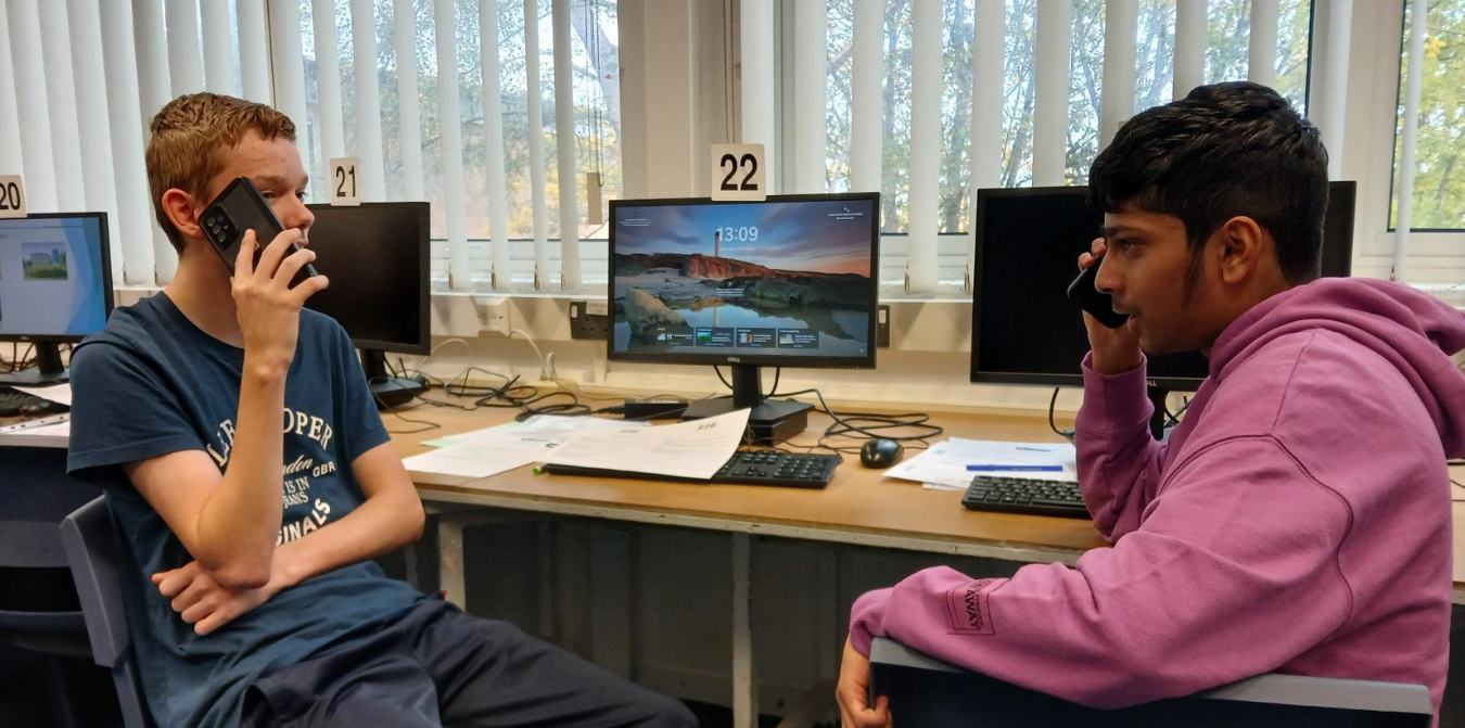 Two people sit at desks in a computer lab, each on a phone. The room has multiple monitors.