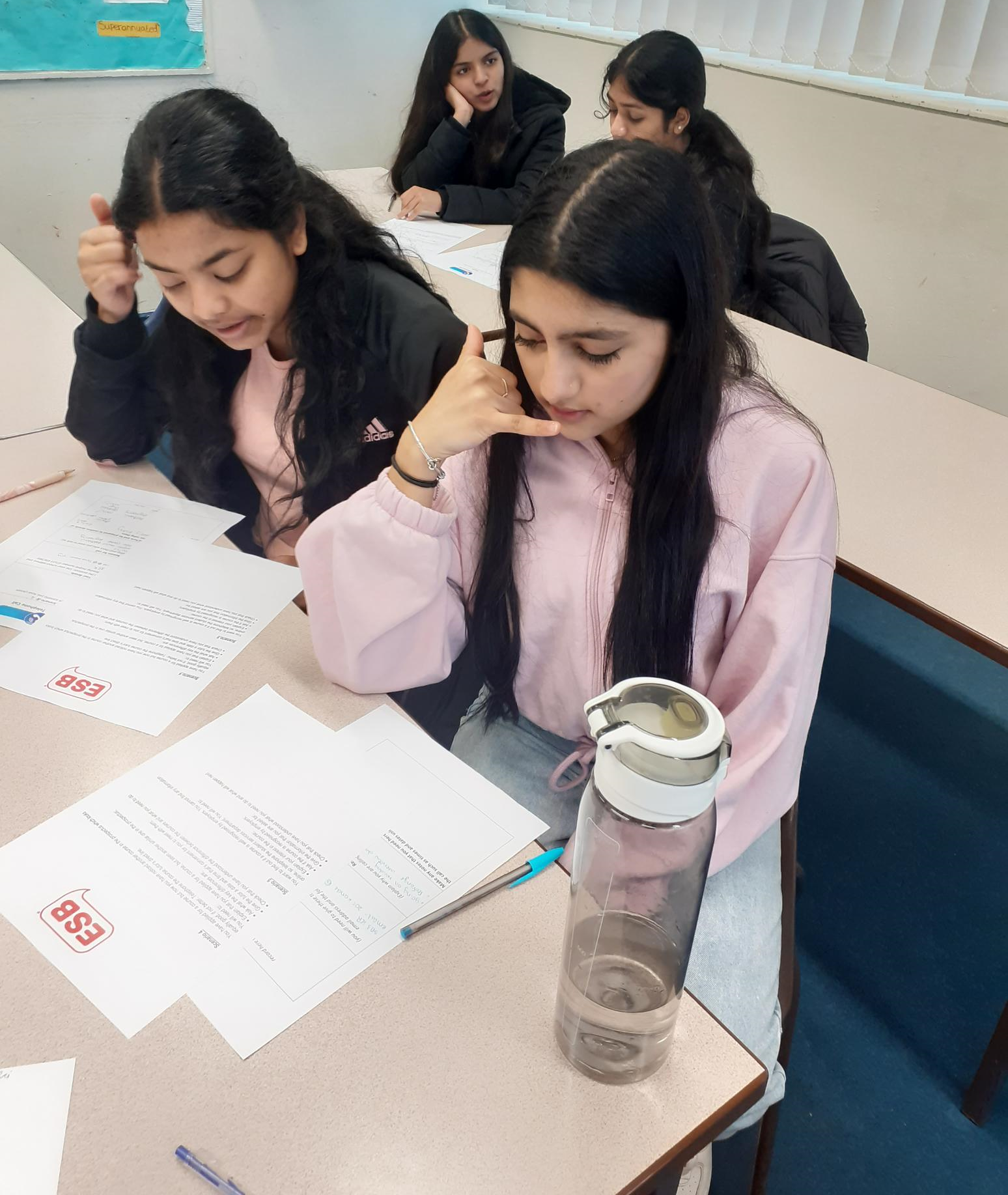 A girl in a pink jacket sits at a table with papers and a water bottle