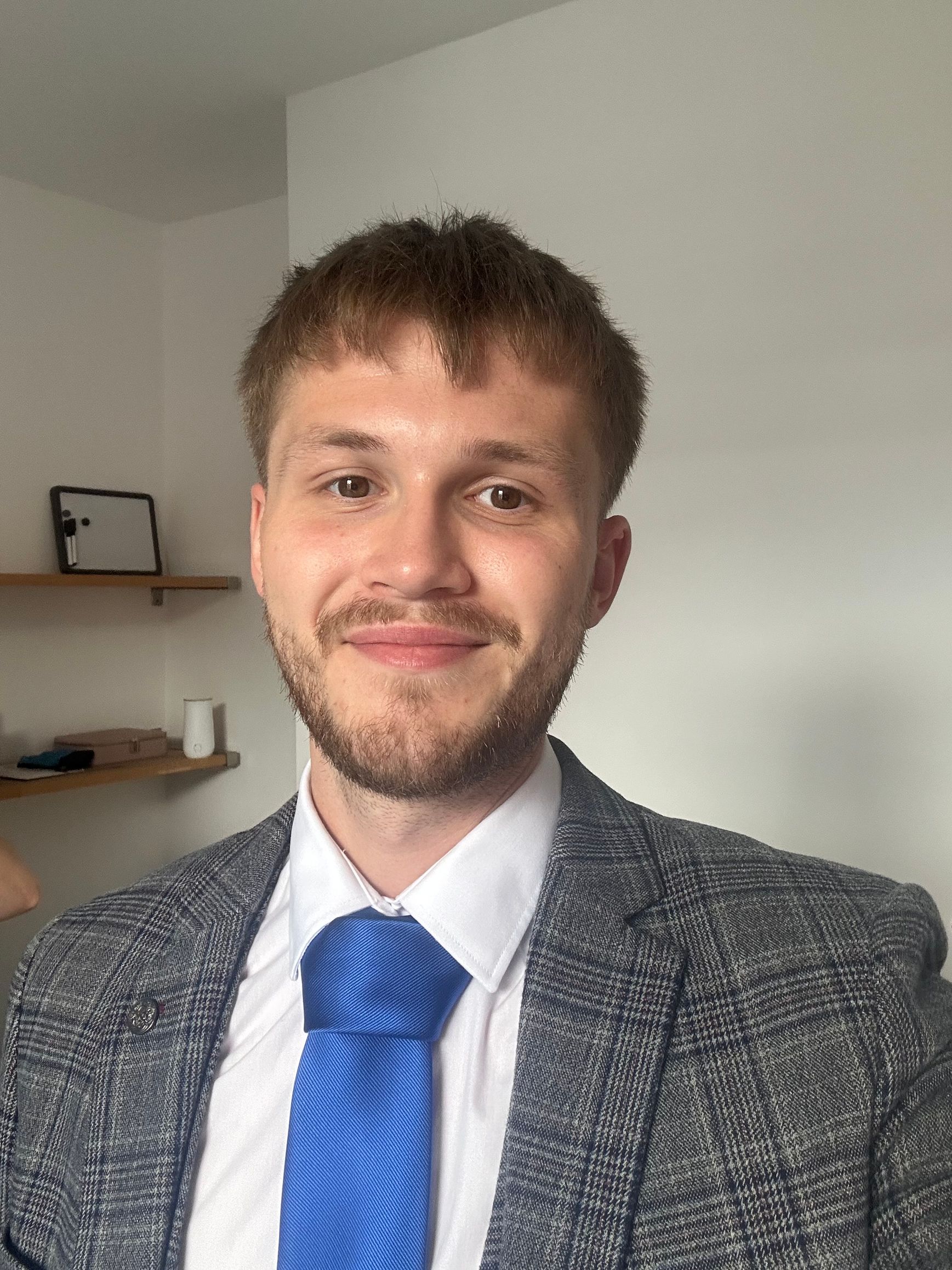 Man in a suit smiles at the camera; blue tie, light gray jacket, white shirt, indoor setting.