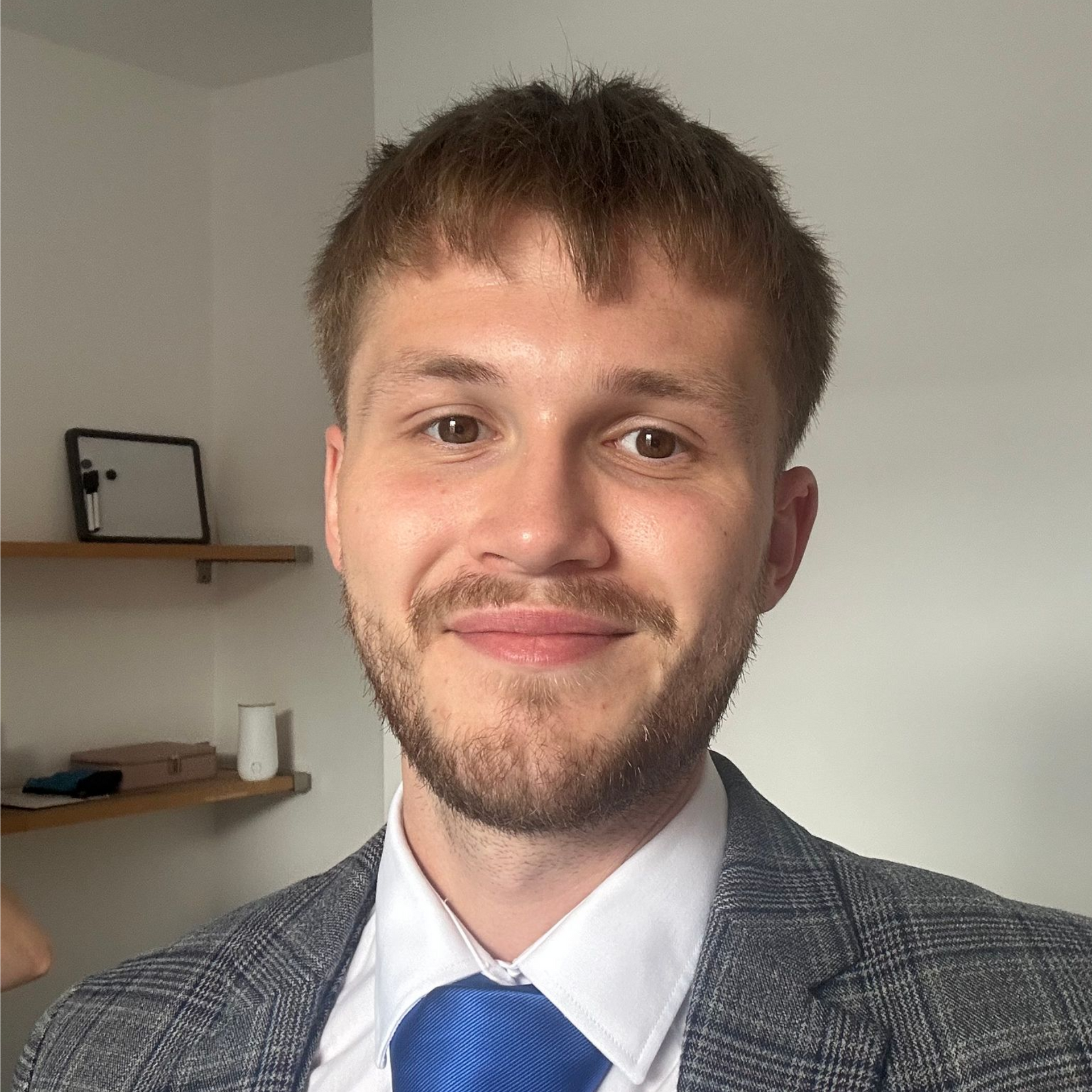 Man in gray blazer and blue tie smiles at the camera. Neutral background, indoors.