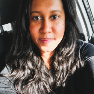 Close-up selfie of a person with long wavy dark hair, wearing a black top, seated in a car