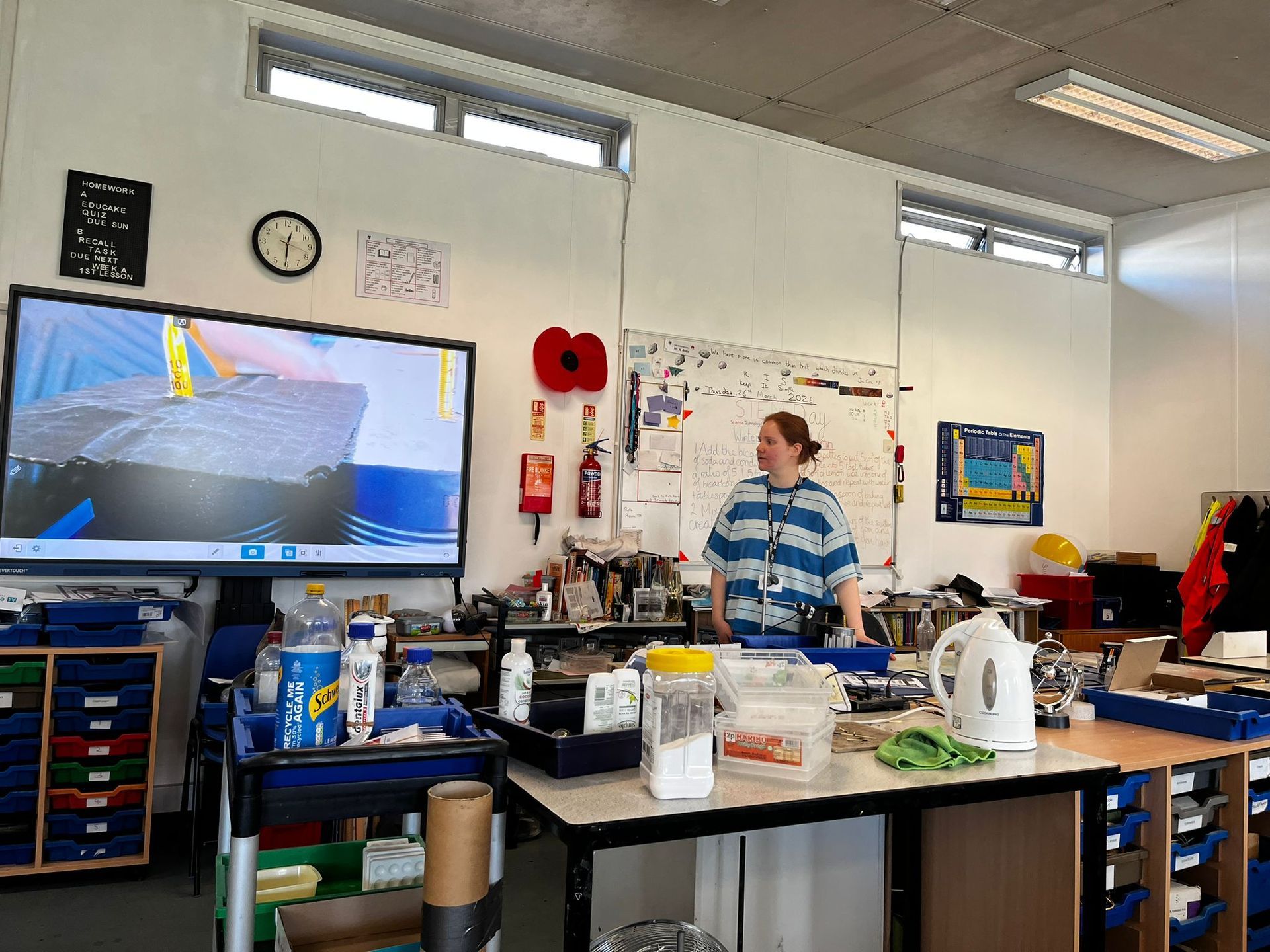 Person standing in a workshop beside a workbench, with tools, shelves, and a wall-mounted monitor nearby