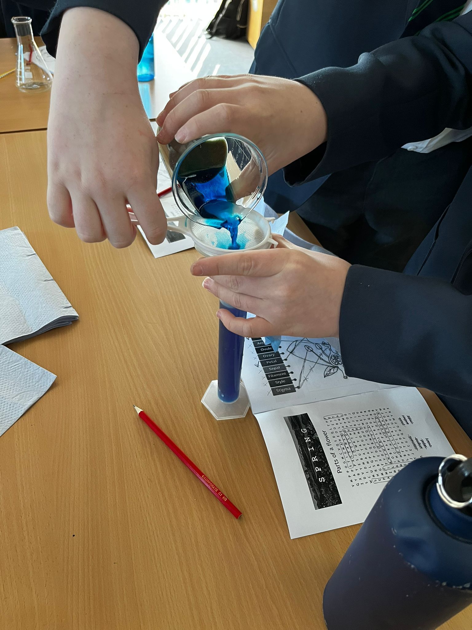 Hands pouring blue liquid into a clear container on a classroom table with papers and a red pencil.