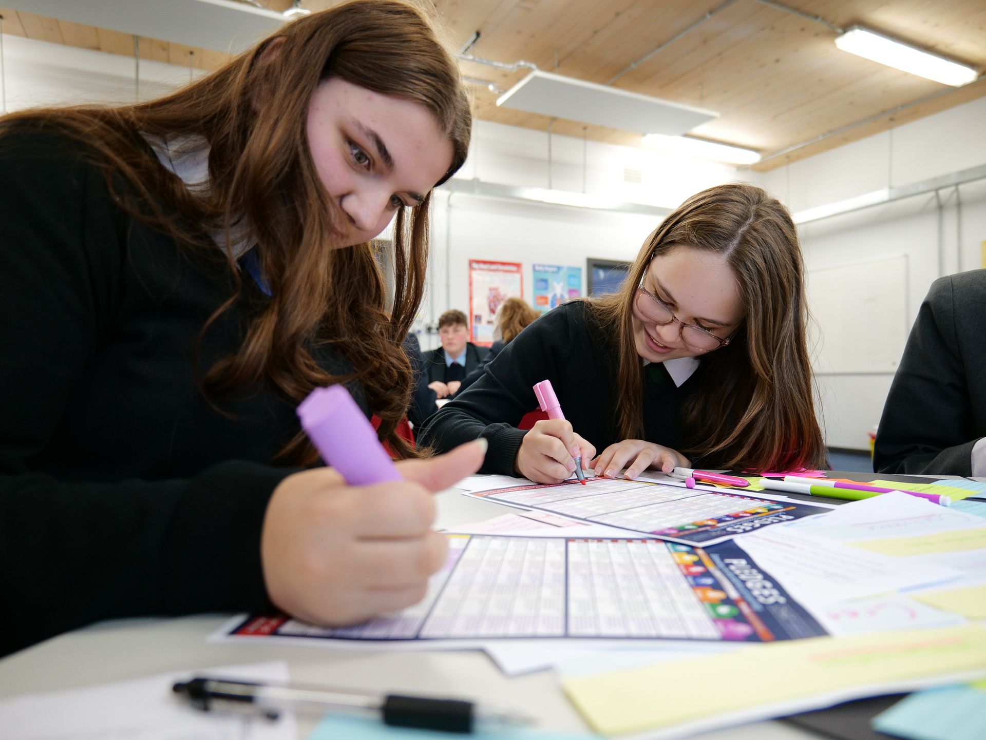 Two girls are sitting at a table in a classroom writing on a piece of paper.