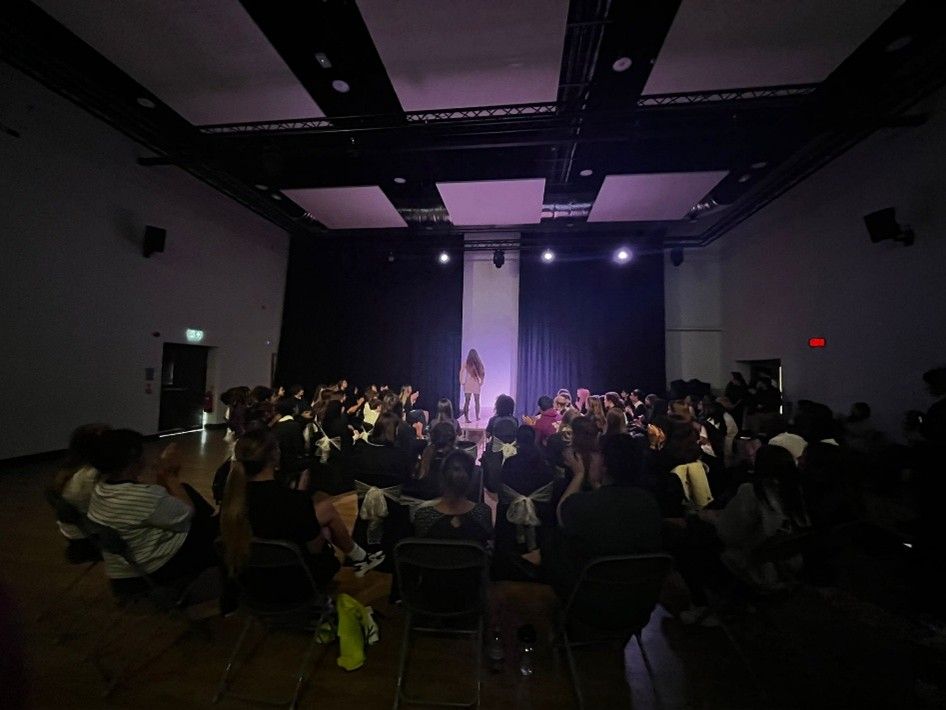 Audience seated in a dimly lit hall facing a stage with purple spotlights