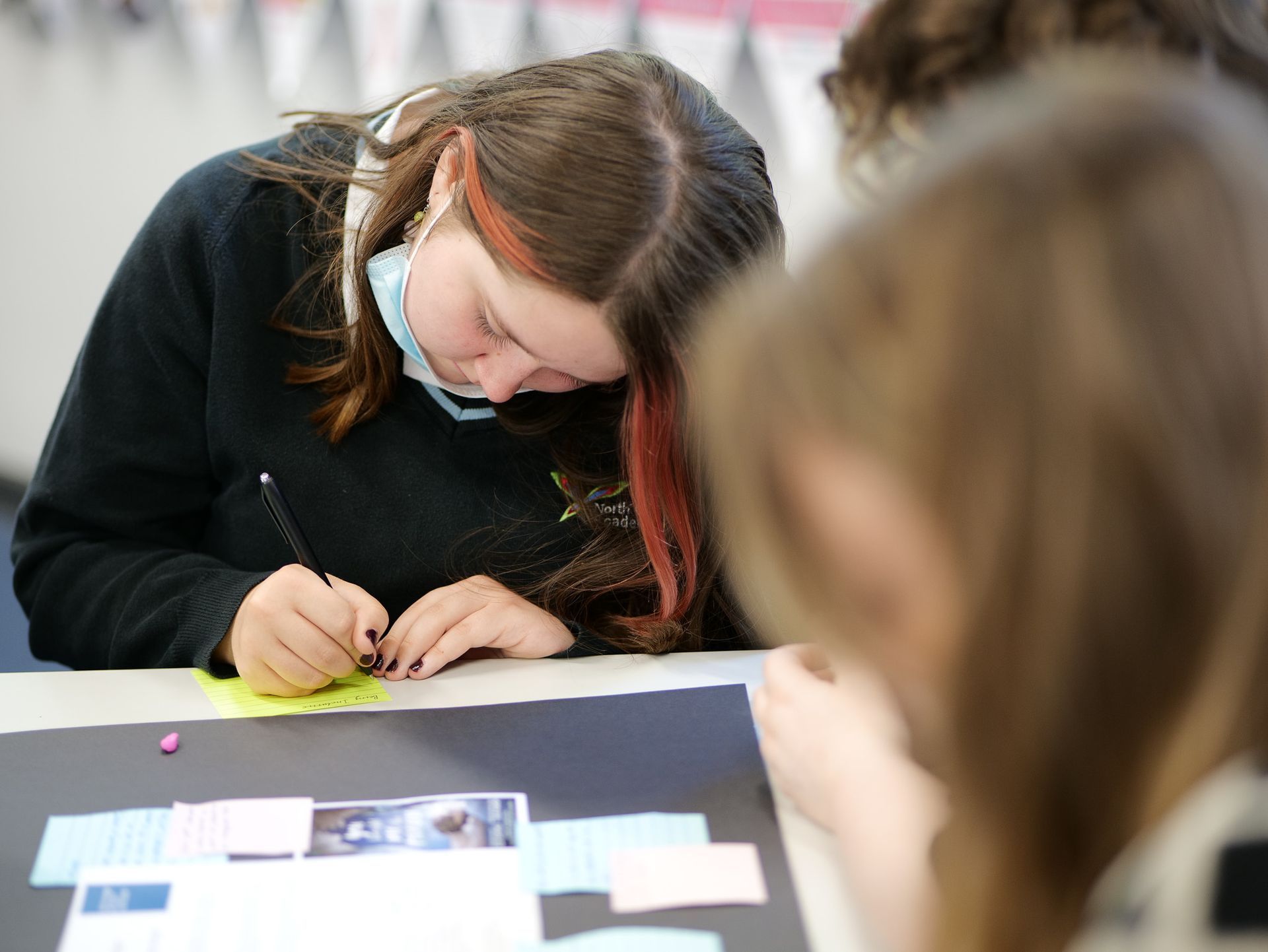Person in black shirt draws with a marker at a table while another person sits blurred in the foreground.