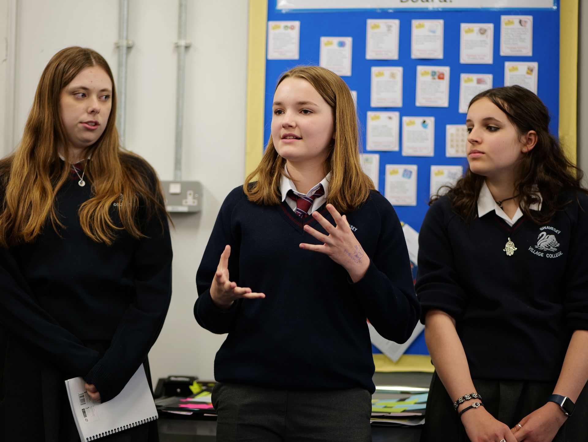 Three girls in school uniforms are standing in front of a blue board
