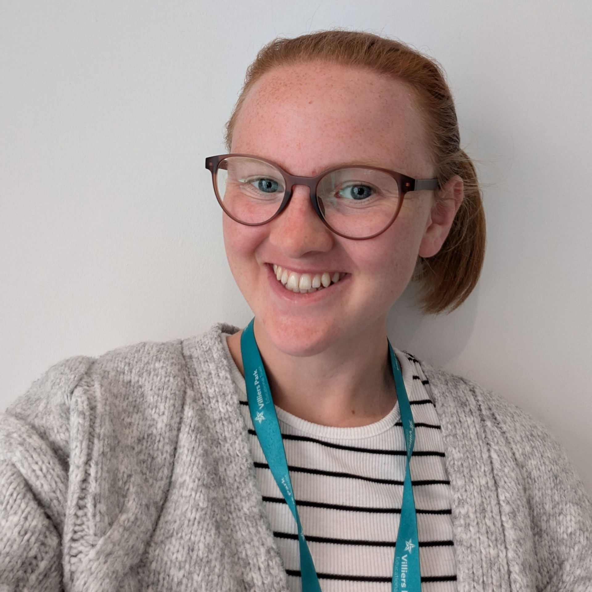Smiling person wearing glasses, striped shirt, gray cardigan, and teal lanyard against a plain white wall