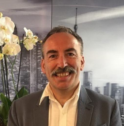 A man with a mustache is smiling in front of a picture of a city.