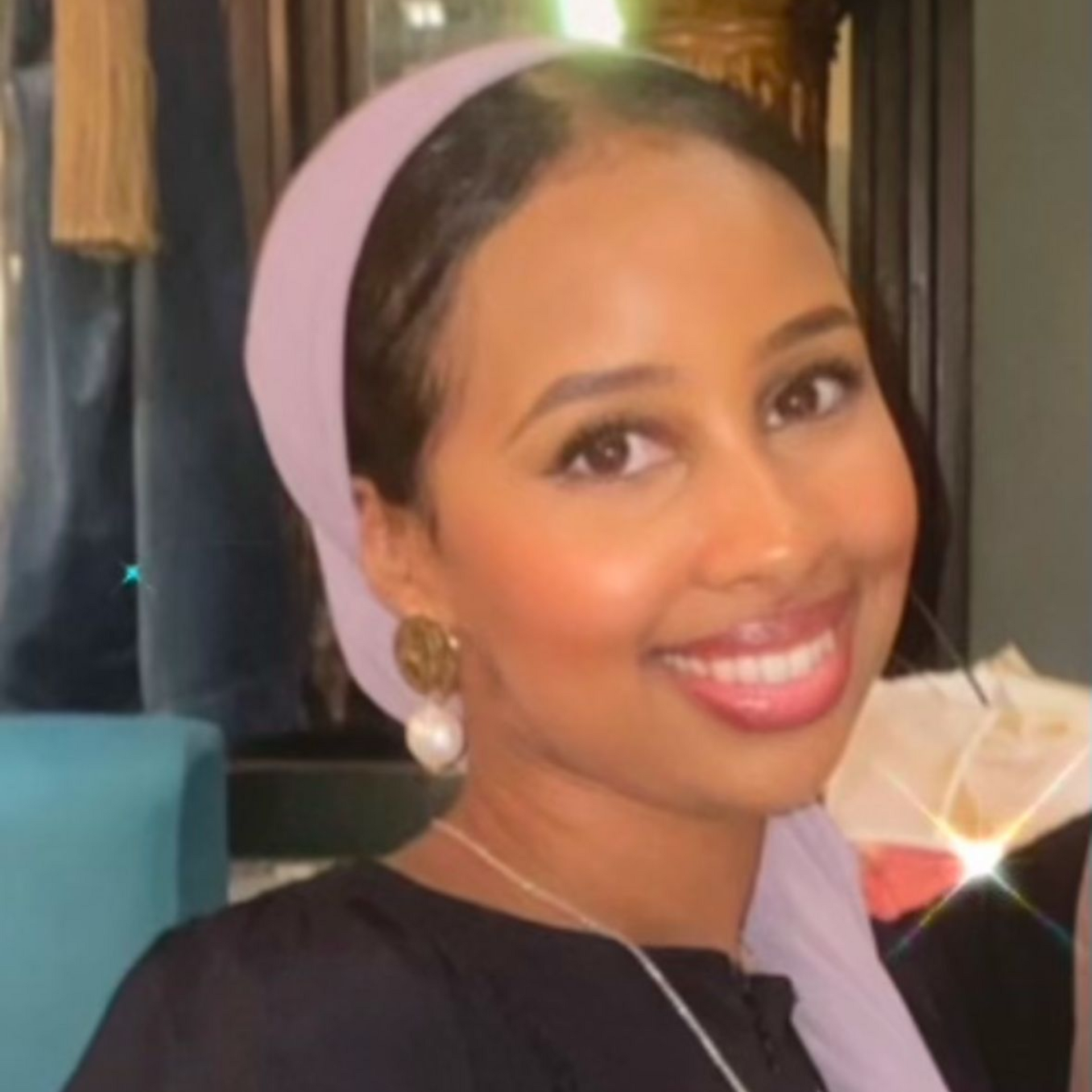 Woman with light skin wearing a light purple headscarf and gold hoop earrings smiling.