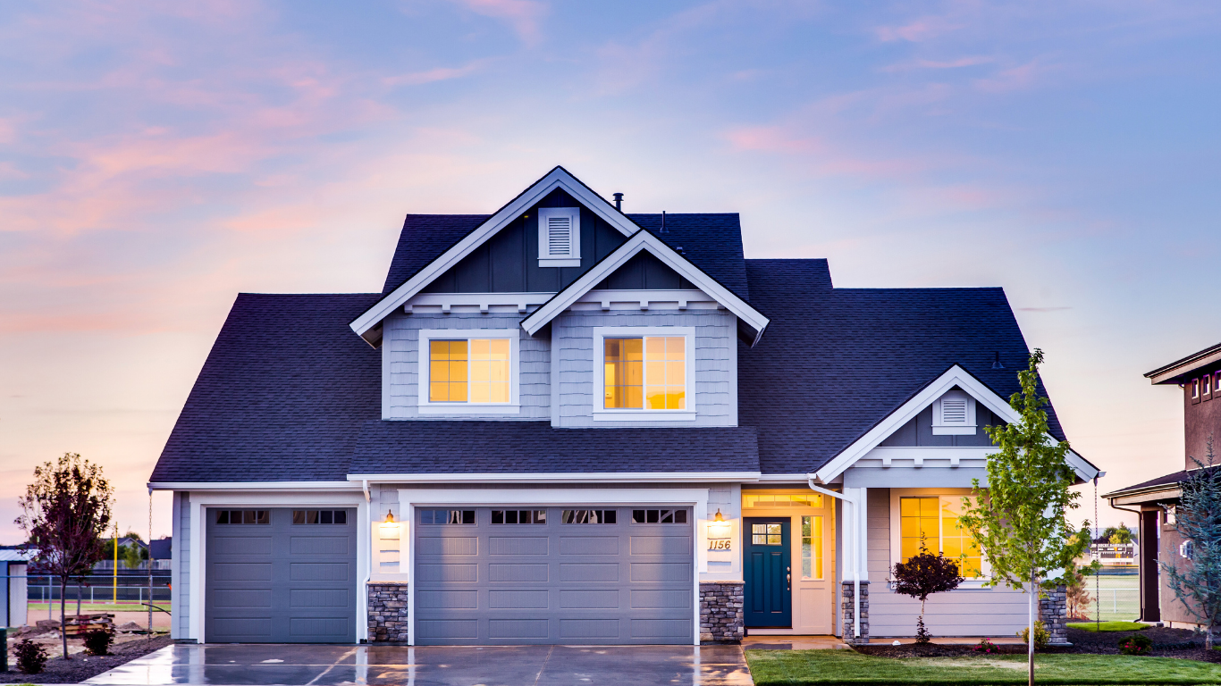 Two-story house with gray exterior, blue door, and a three-car garage, set against a pastel sunset sky.