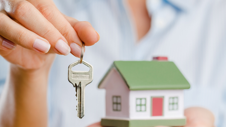 Hand holding house key above a miniature house model.