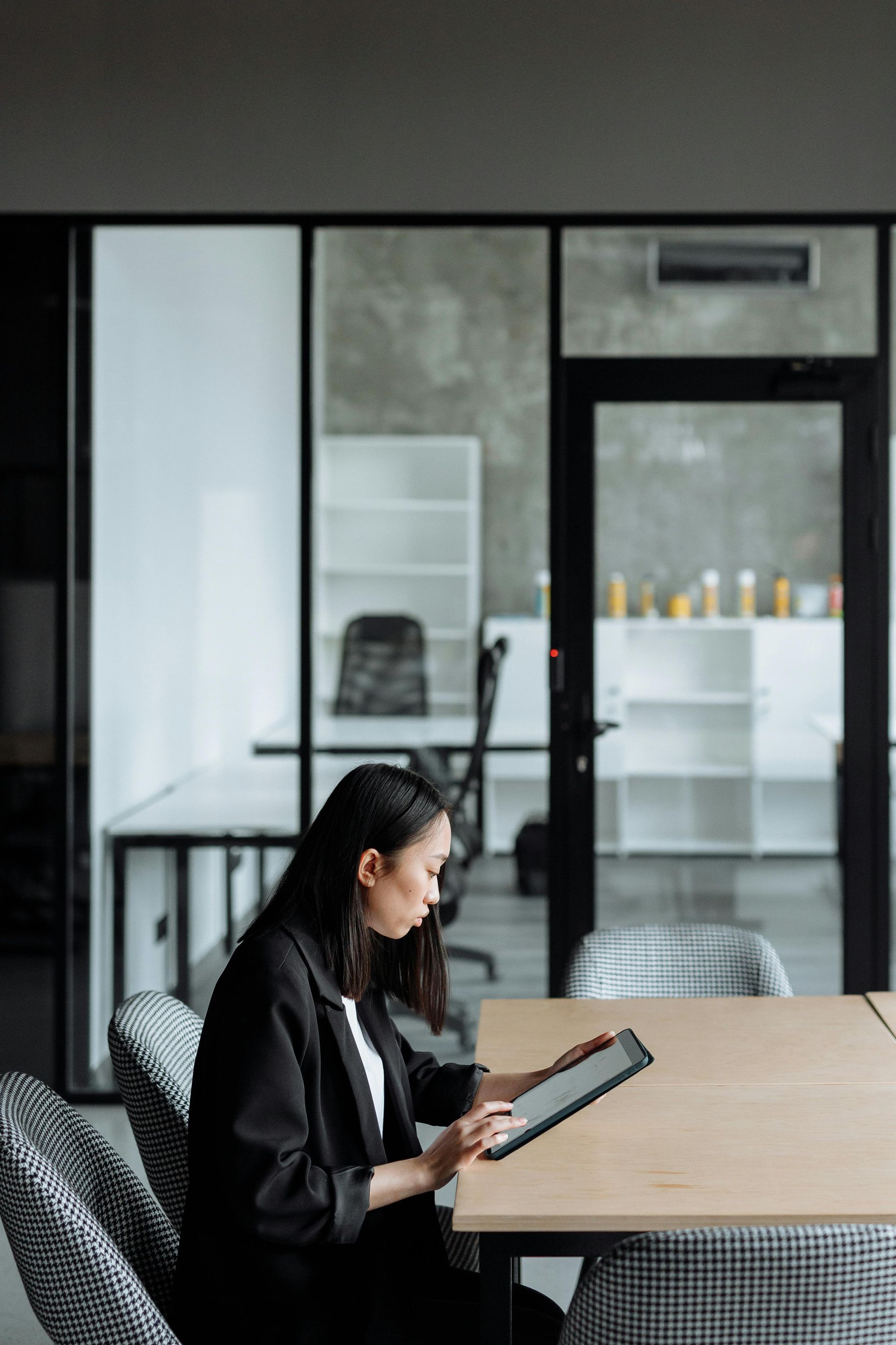 Woman in black blazer at table, using tablet in office setting.