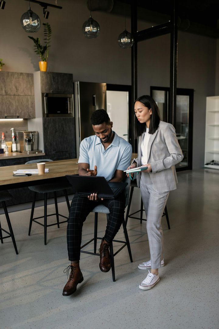 Man seated, working on a laptop, with a woman beside him reviewing documents. Modern office with dark furniture.