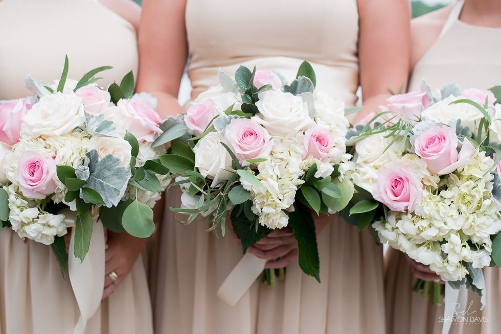 Three women hold pastel pink and white floral bouquets with greenery, dressed in blush dresses.