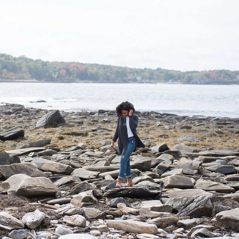 Person standing on rocky shoreline by a calm lake, wearing a dark jacket and jeans.