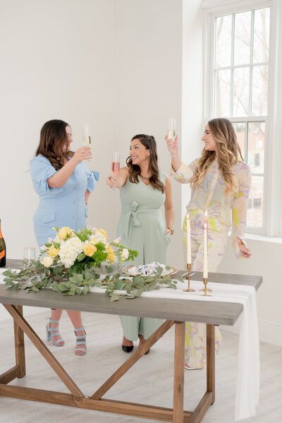 Three women toast with drinks beside a flower-decorated table in a bright room.