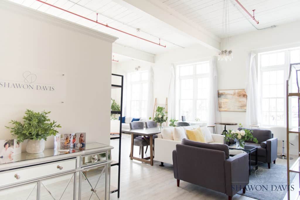 Bright living room with white walls, large windows, a dark sofa, and a console table with plants and decor.